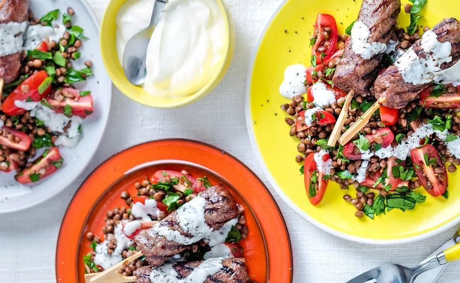 Grilled skewers with lentil salad and yogurt sauce on colorful plates.