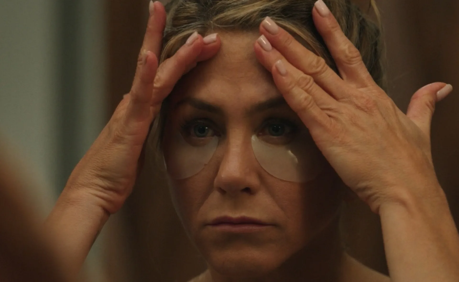 Woman wearing under-eye patches, pressing palms to forehead, looking in mirror.