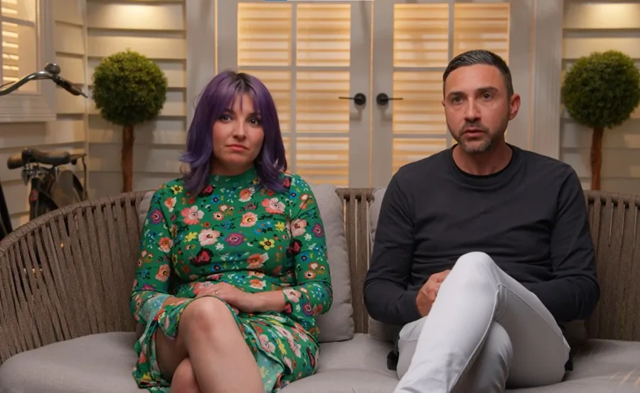 Two people seated indoors; one in a floral dress with purple hair, the other in a black shirt and white pants.