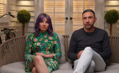 Two people seated indoors; one in a floral dress with purple hair, the other in a black shirt and white pants.