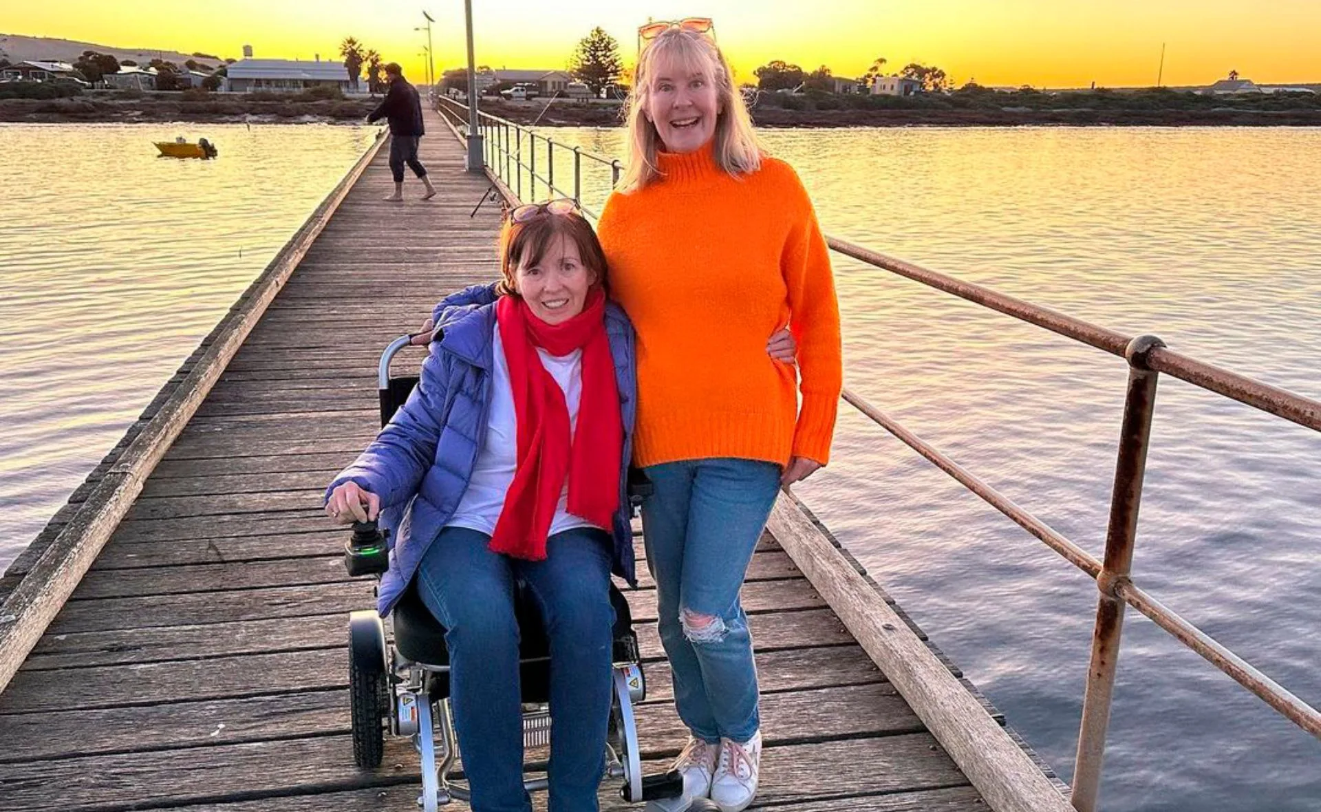 Two women smiling on a wooden pier at sunset, one in a wheelchair, next to calm water.