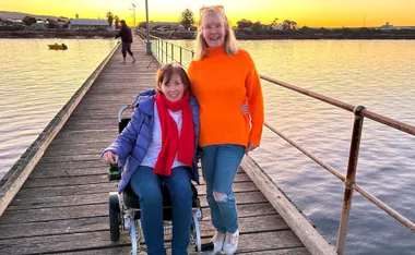 Two women smiling on a wooden pier at sunset, one in a wheelchair, next to calm water.