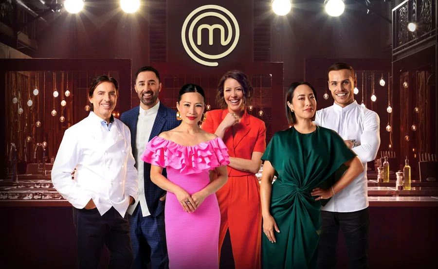 "MasterChef judges and contestants standing together in the iconic kitchen set, with the MasterChef logo in the background."