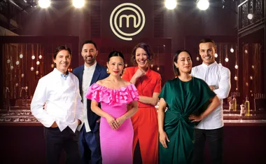 "MasterChef judges and contestants standing together in the iconic kitchen set, with the MasterChef logo in the background."