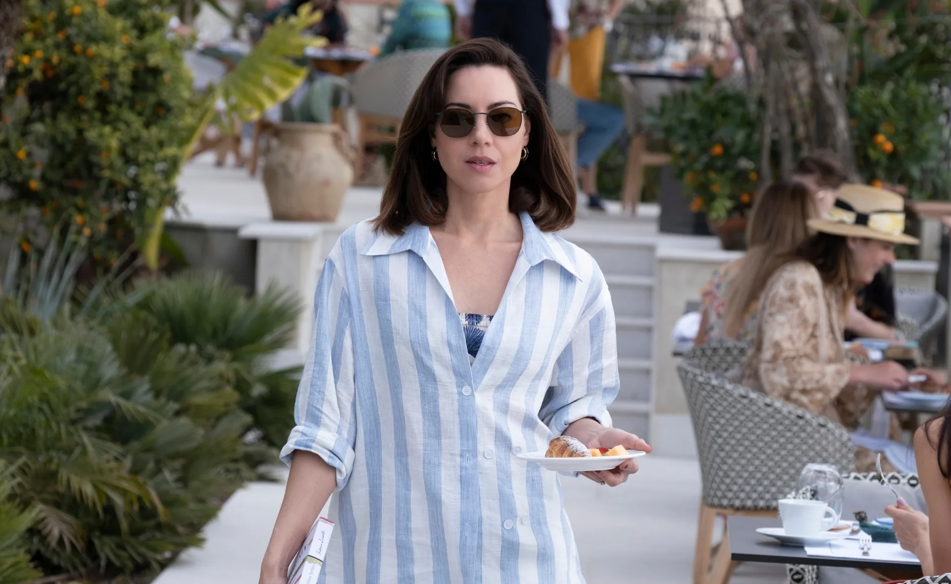 Woman in sunglasses and striped shirt holds plate in outdoor cafe setting.