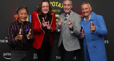 Four men smiling and gesturing with finger guns at a red carpet event, dressed in colorful suits.