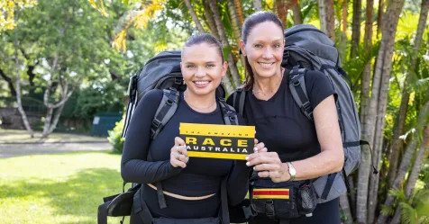 Two women with backpacks holding a "Race Australia" card in a lush, green outdoor setting, preparing for the challenge.