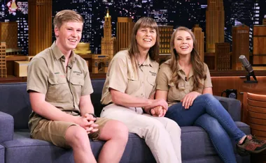 A family of three wearing khaki shirts sits on a couch, smiling during a TV interview against a backdrop of a city skyline.