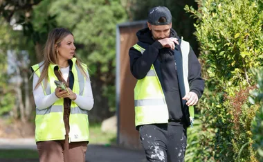 Two people in high-visibility vests walk outdoors, one using a smartphone, the other with a hand near the face.