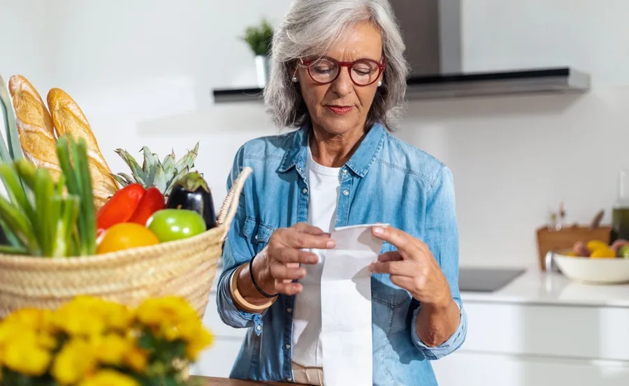 Elderly woman in a kitchen reviewing a grocery receipt beside a basket of vegetables and bread.