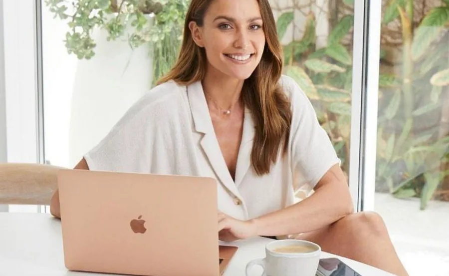 Smiling woman sitting at a table with a laptop and coffee, indoor plants in the background.
