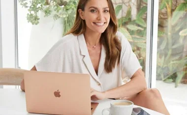 Smiling woman sitting at a table with a laptop and coffee, indoor plants in the background.