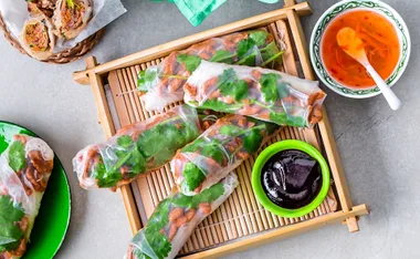 Char siu pork rice paper rolls on a bamboo tray with dipping sauces and fresh herbs, accompanied by a bowl of sauce.