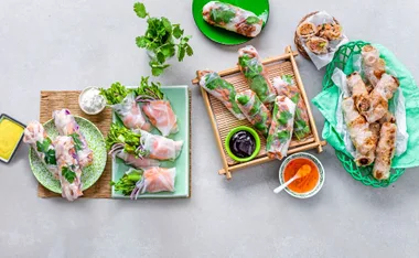 Assorted rice paper rolls with fresh herbs and dipping sauces on a light background.