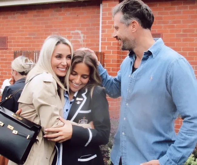 A woman and a girl hug while a man smiles at them, standing outside in front of a brick wall.