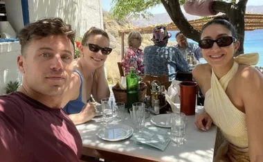 Group of three people smiling at an outdoor restaurant table, with a scenic view in the background.