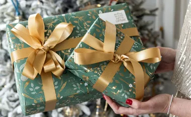 Two gifts wrapped in green floral paper with gold ribbons held near a Christmas tree.