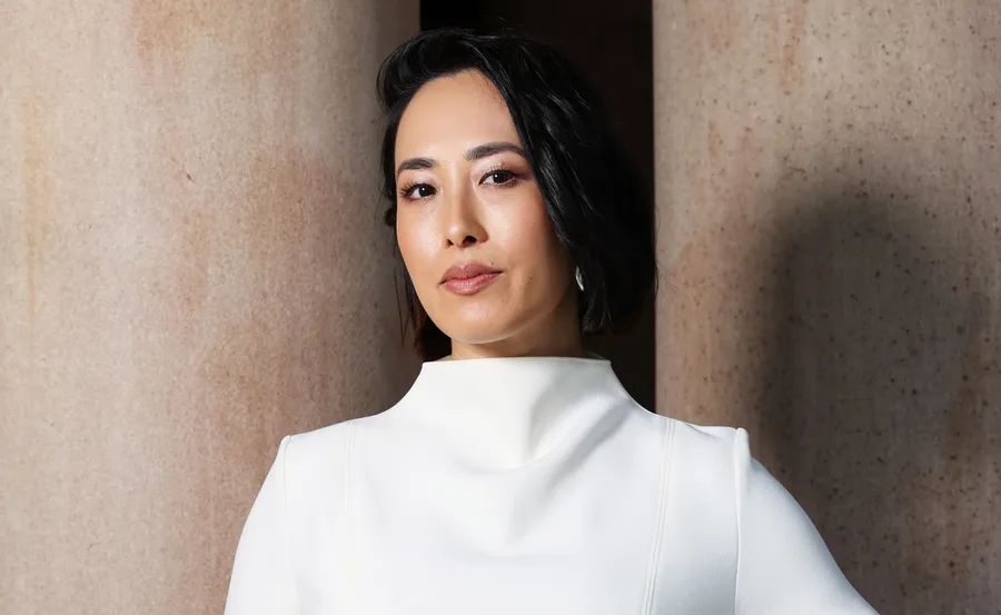 A person with short black hair wearing a white outfit, standing in front of large columns.