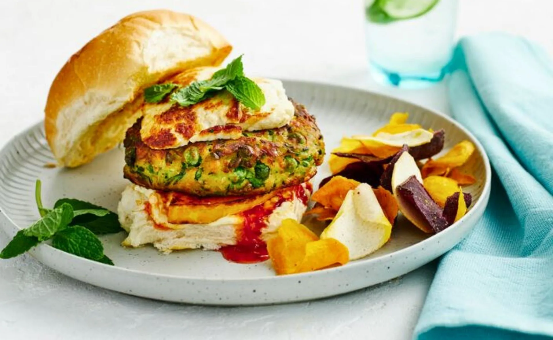Vegan burger with cheese and mint on a bun, served with mixed vegetable chips on a plate.