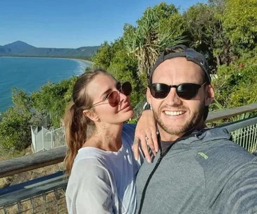 A couple taking a selfie with a scenic coastal landscape in the background under a clear blue sky.