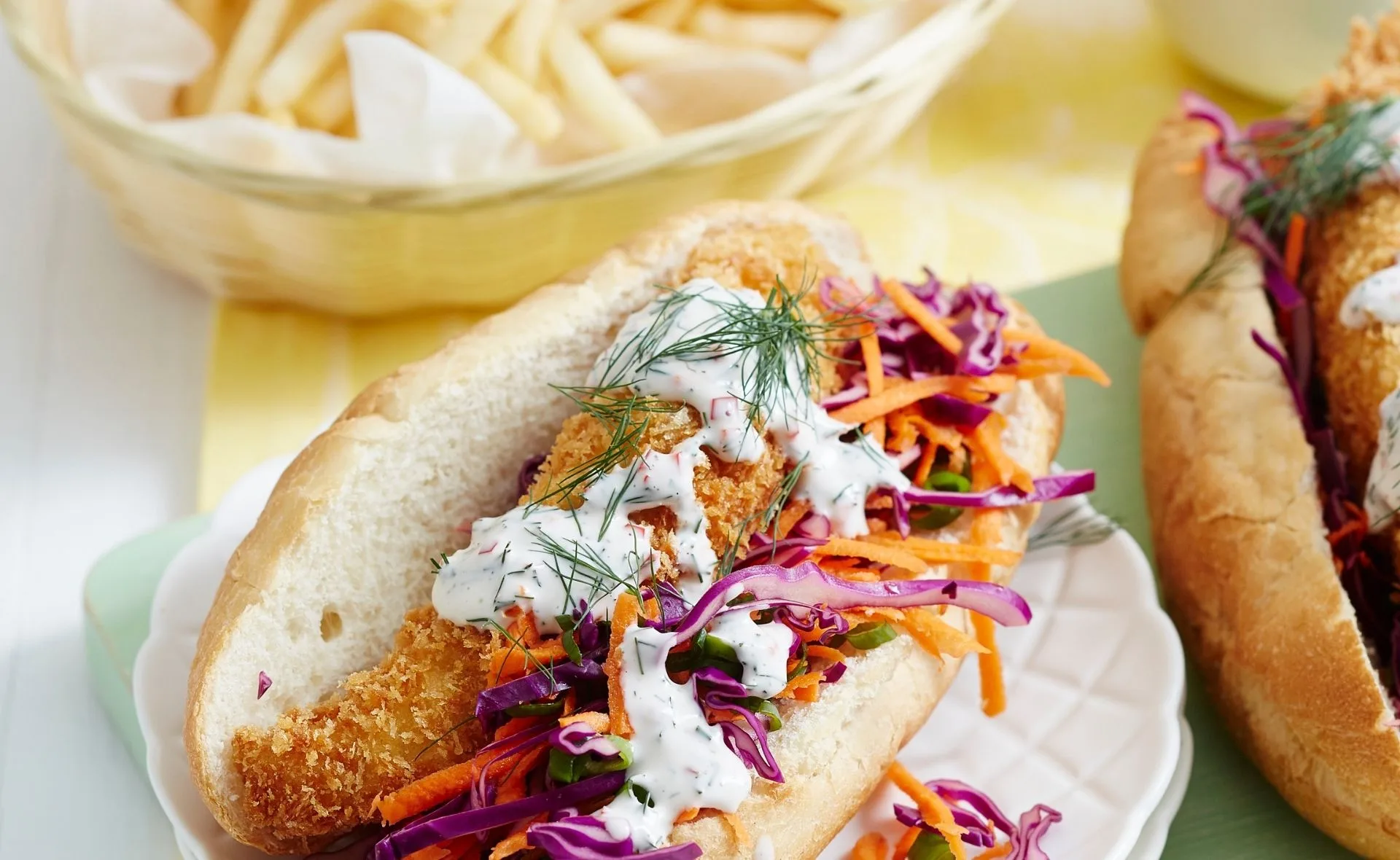 Crumbed fish burger with carrots, purple cabbage, and dill sauce on a roll, fries in background.