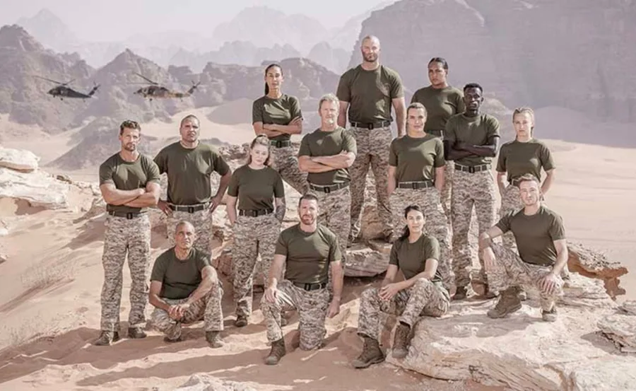 A group of 14 people in military uniforms posing on a rocky desert landscape with helicopters in the background.