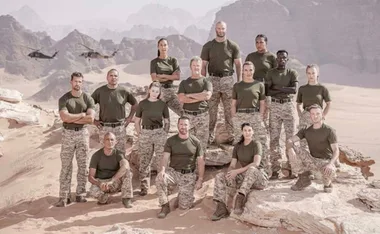 A group of 14 people in military uniforms posing on a rocky desert landscape with helicopters in the background.