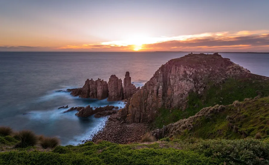 Sunset over the ocean cliffs at Phillip Island, featuring rugged rock formations and lush greenery.