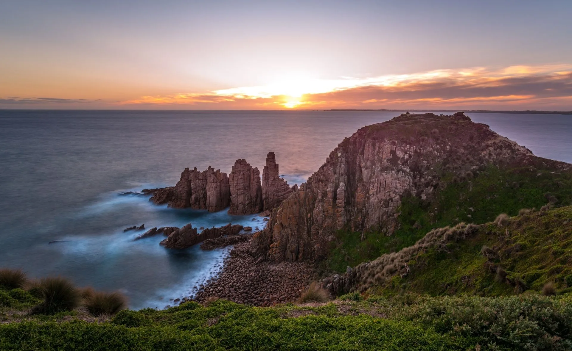 Sunset over the ocean cliffs at Phillip Island, featuring rugged rock formations and lush greenery.