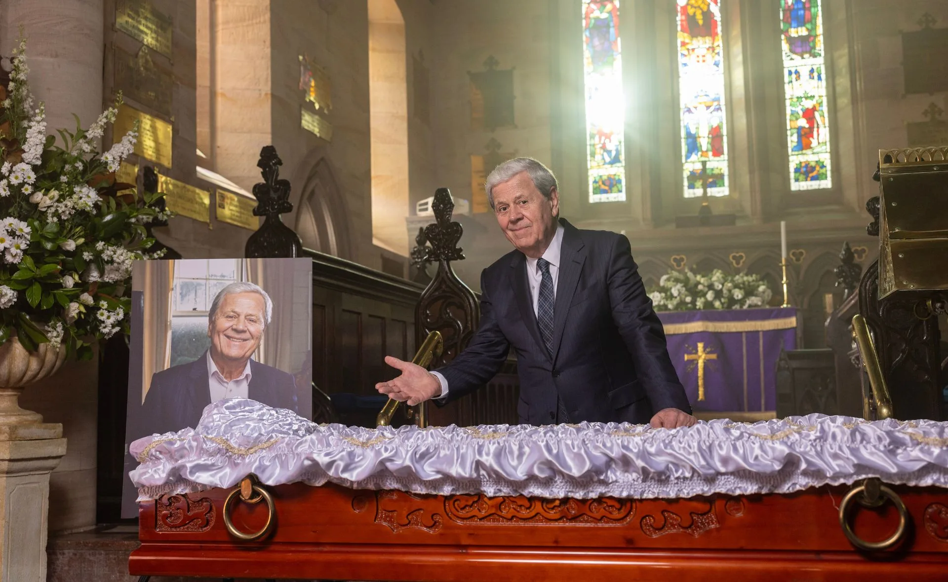 A man stands beside a portrait at a decorated coffin in a church, with stained glass windows in the background.