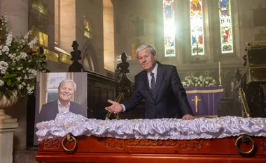A man stands beside a portrait at a decorated coffin in a church, with stained glass windows in the background.
