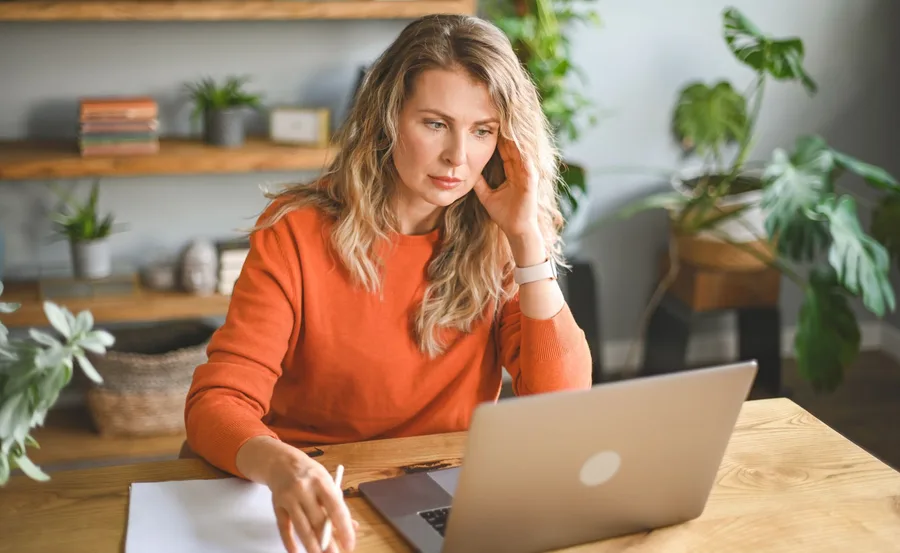 A woman in an orange sweater looks stressed while working on a laptop at a wooden desk with plants in the background.