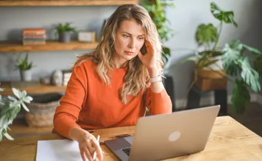 A woman in an orange sweater looks stressed while working on a laptop at a wooden desk with plants in the background.