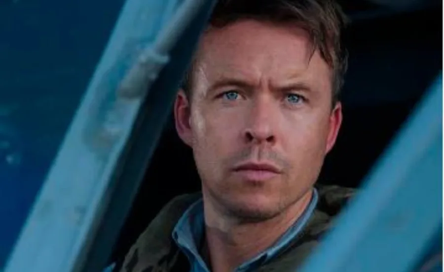 A man with short hair and blue eyes looking out of a window with a focused expression.