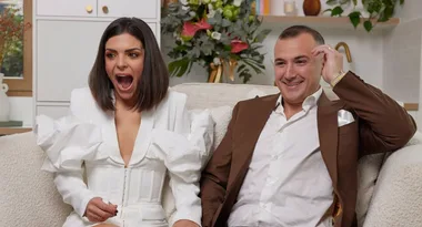 Two people sit on a couch, one looks surprised, the other amused; both dressed formally.