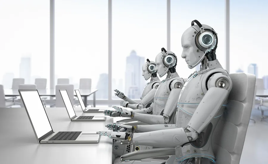 Robots in an office setting, seated in a row, typing on laptops, suggesting automation or AI in the workplace.