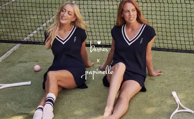 Two women sit on a tennis court in matching navy dresses, with rackets and a pink ball nearby.