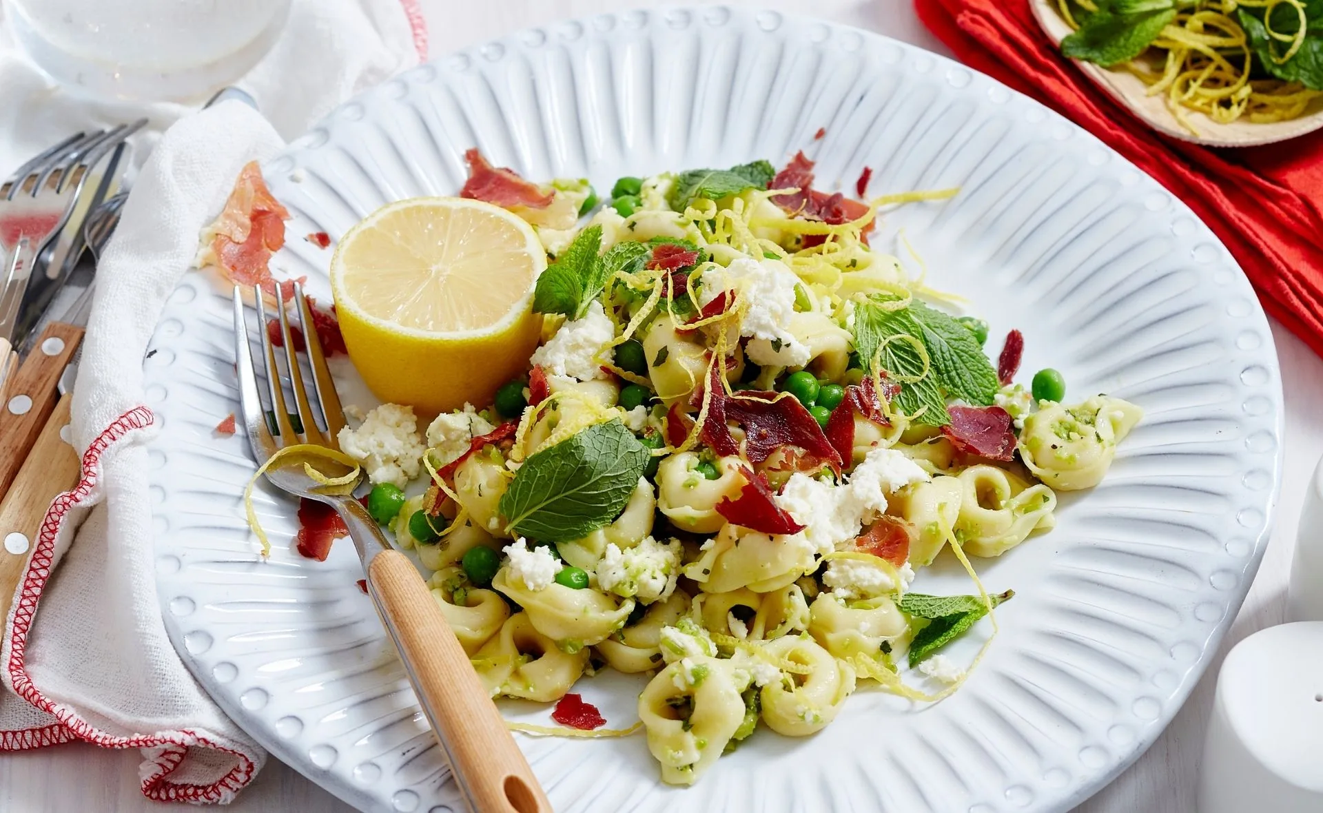 Plate of tortellini with prosciutto, peas, ricotta, mint, lemon zest, and a lemon half on a white plate.