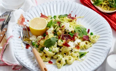 Plate of tortellini with prosciutto, peas, ricotta, mint, lemon zest, and a lemon half on a white plate.