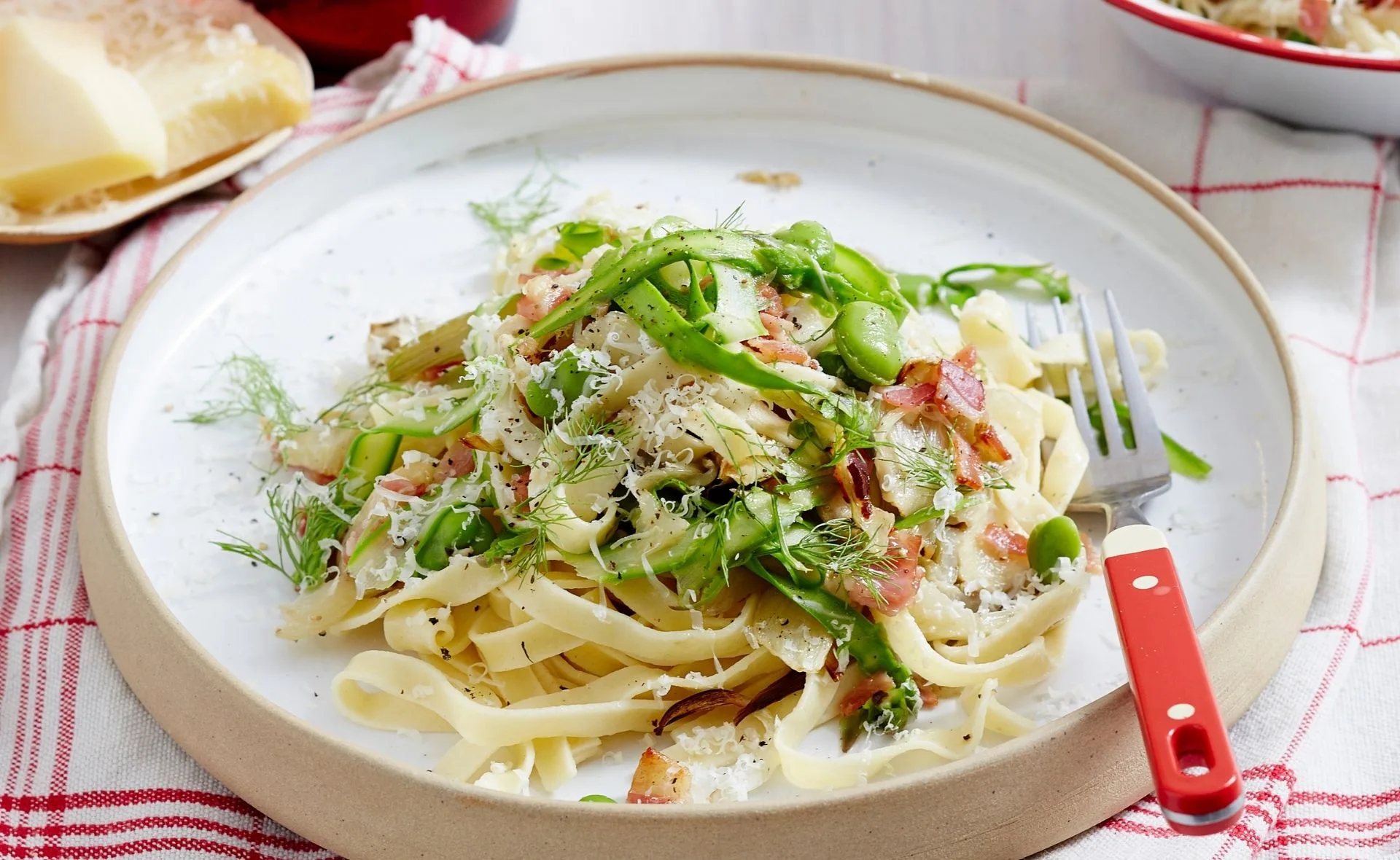 Plate of fettuccine with sliced asparagus, broad beans, and fennel, garnished with grated cheese and a red-handled fork.