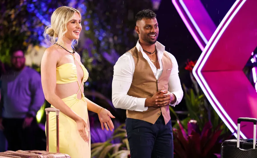 Two people stand with luggage under bright lights on a reality show set, smiling and engaging with the scene.