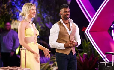 Two people stand with luggage under bright lights on a reality show set, smiling and engaging with the scene.