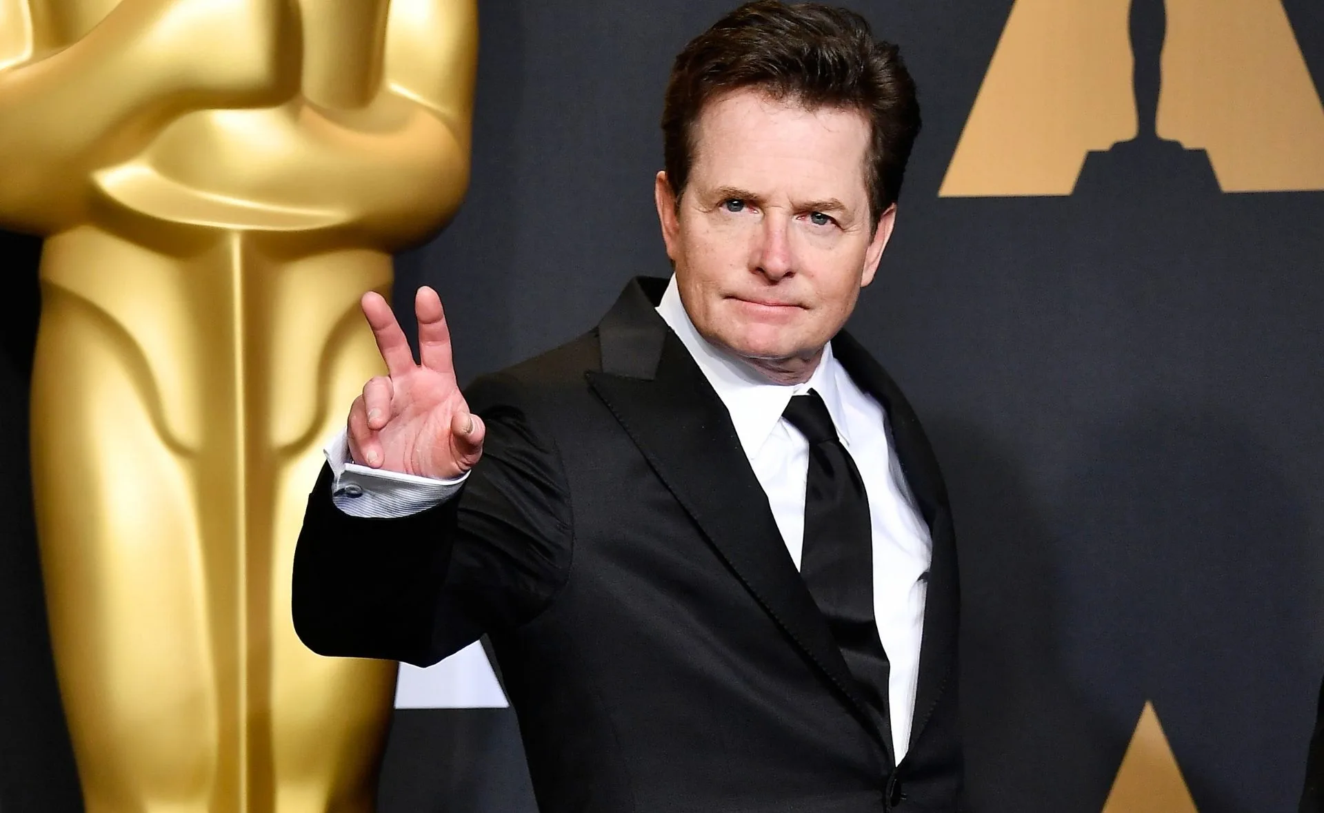 A person in a suit making a peace sign gesture, standing in front of a large golden statue.