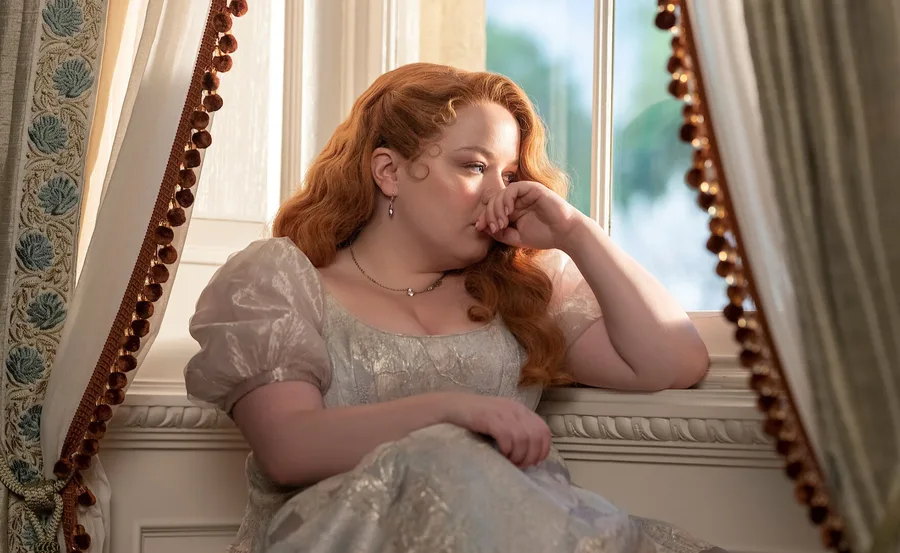 A woman with red hair in a period costume looks thoughtful by a window decorated with ornate curtains.