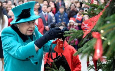 Elegant elderly woman in teal attire hangs a decoration on a Christmas tree before a crowd.