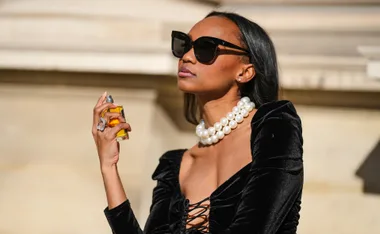 Woman in black dress and pearl necklace, holding perfume bottle, wearing sunglasses.