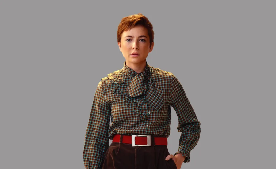 A person wearing a plaid shirt with puff sleeves and a red belt, standing against a gray background.
