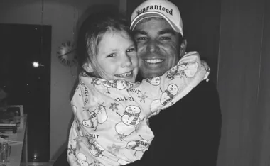 Father and daughter share a joyful hug, with the girl wearing snowman pajamas and the father in a cap.