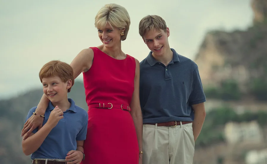 A woman in a bright red dress stands smiling with two young boys in casual shirts, set against a blurred outdoor backdrop.