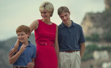 A woman in a bright red dress stands smiling with two young boys in casual shirts, set against a blurred outdoor backdrop.